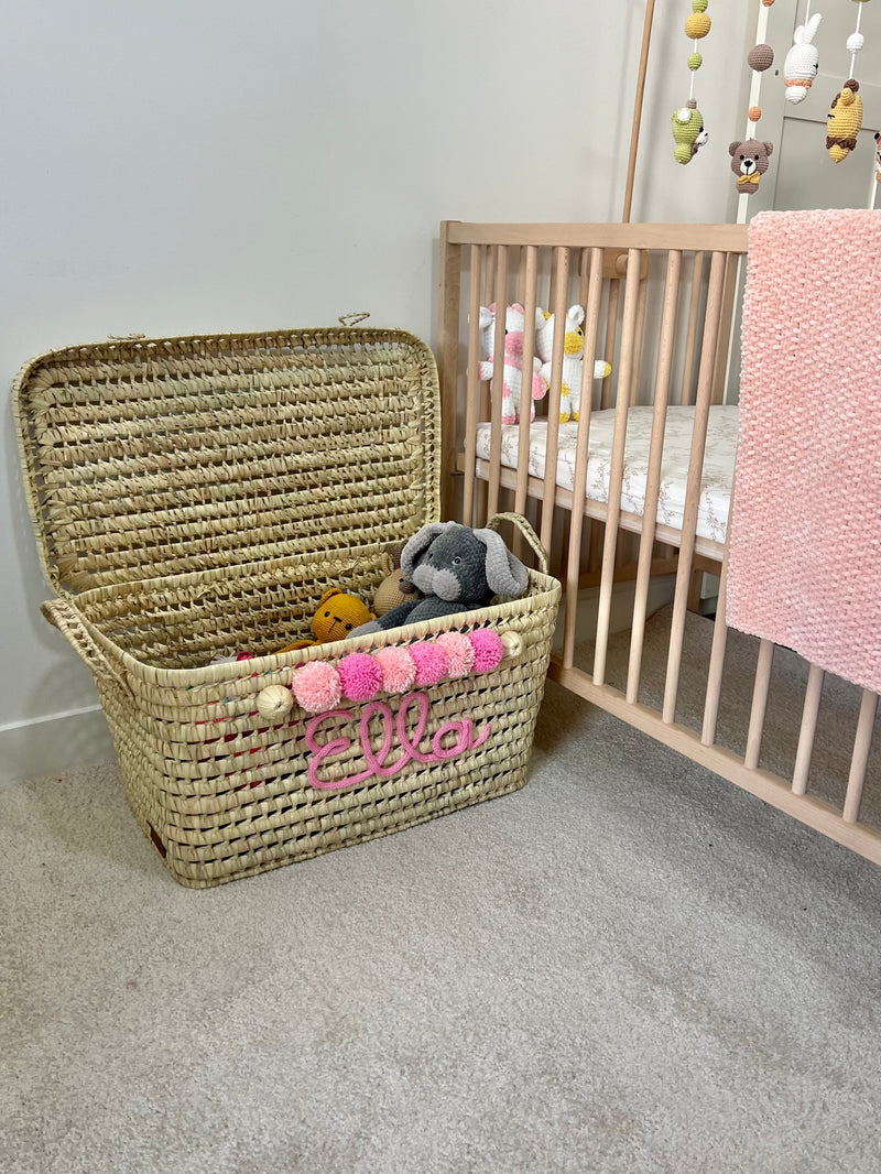 Coffre à jouets en osier ouvert avec peluches, personnalisé au prénom Ella en tricotin rose et pompons roses dans une chambre de bébé