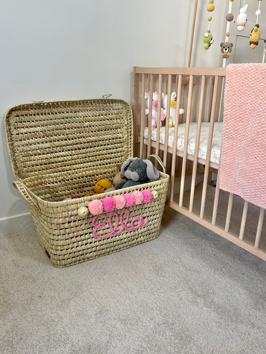 Coffre à jouets en osier ouvert avec peluches, personnalisé au prénom Ella en tricotin rose et pompons roses dans une chambre de bébé