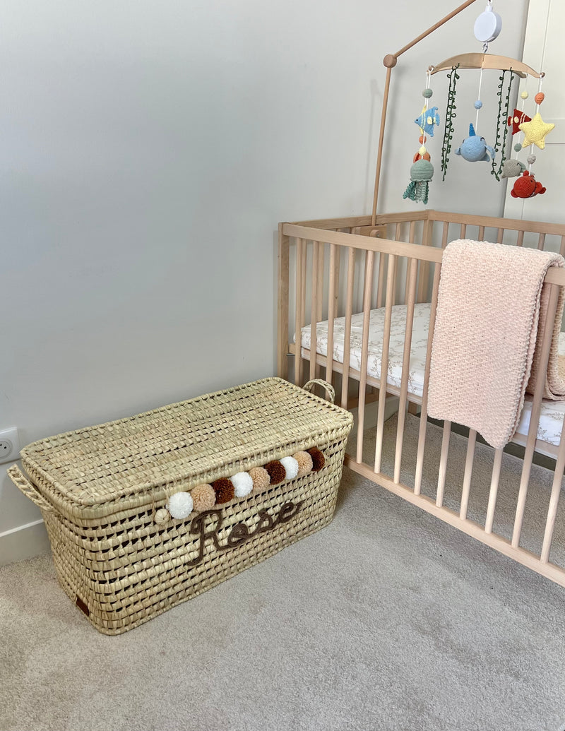 Malle à jouets personnalisée en osier avec prénom en tricotin marron et pompons beiges, blancs et marron, près d'un lit de bébé