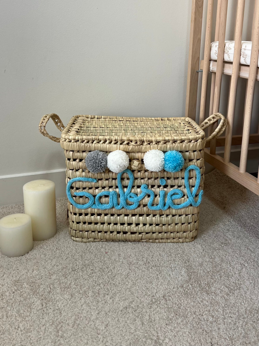 Malle à jouets en osier personnalisée avec prénom bleu et pompons bleus, vue dans une chambre d'enfant