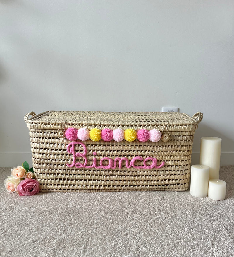Malle en osier avec prénom rose en tricotin et pompons roses et jaunes, décoration de chambre bébé
