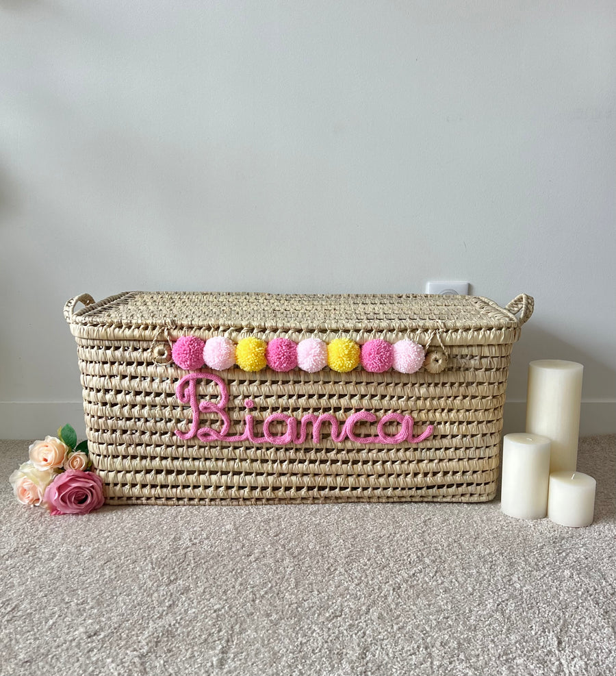 Malle en osier avec prénom rose en tricotin et pompons roses et jaunes, décoration de chambre bébé