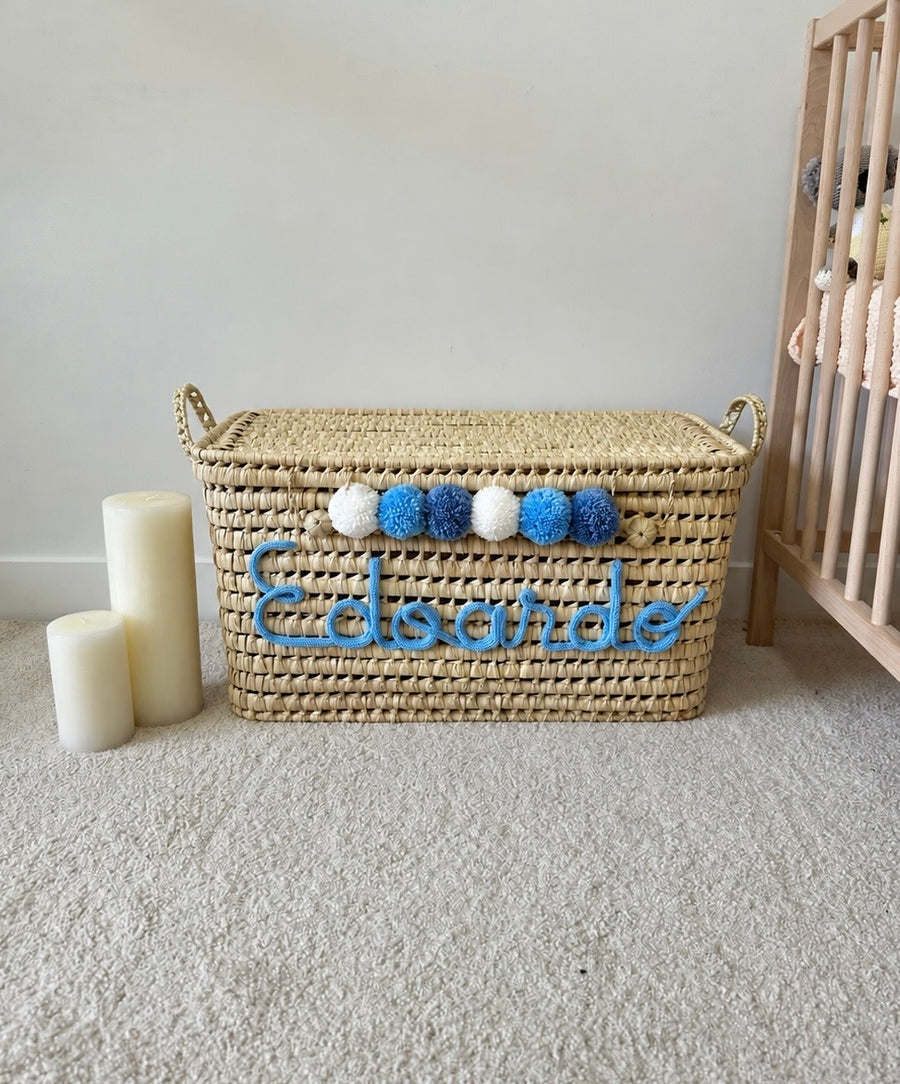 Coffre à jouets en osier personnalisé avec prénom en tricotin bleu et pompons bleus et blancs