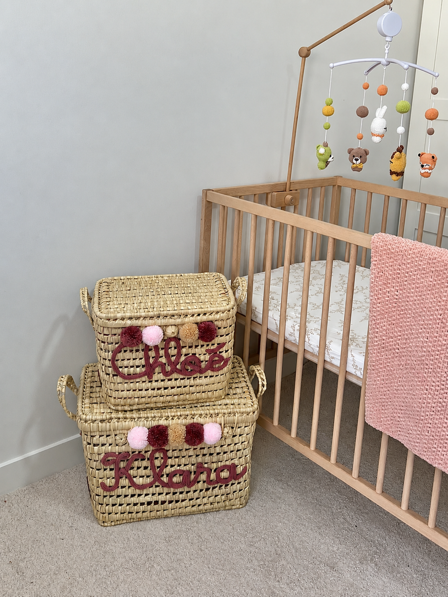 Coffre à jouets personnalisé en osier avec prénom rose et pompons roses, près d'un lit de bébé